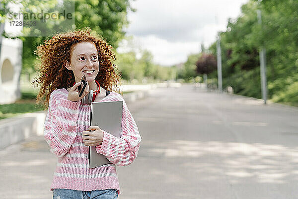 Lächelnde junge Frau mit lockigem Haar  die im Park über einen Lautsprecher mit ihrem Smartphone spricht und dabei wegschaut
