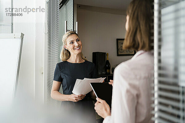 Lächelnde Unternehmerin  die eine Kollegin anschaut  während sie an der Tür im Büro diskutiert