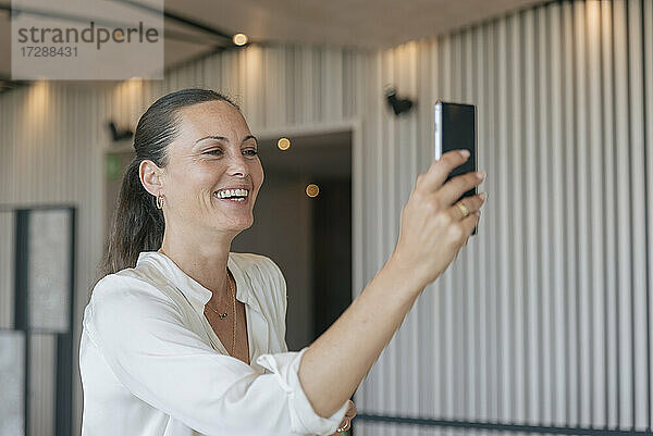 Fröhliche reife Geschäftsfrau bei einem Videoanruf über ein Smartphone in der Lobby