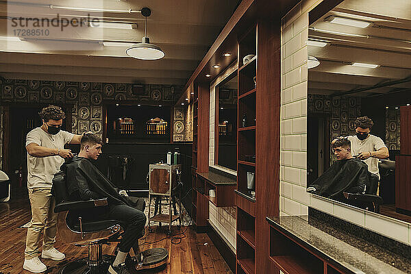 Junger Mann erhält einen Haarschnitt von einem männlichen Friseur in einem Salon während der COVID-19-Krise