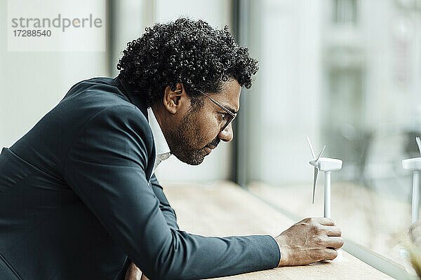 Männlicher Fachmann  der sich auf den Schreibtisch stützt  während er das Modell einer Windkraftanlage im Büro untersucht