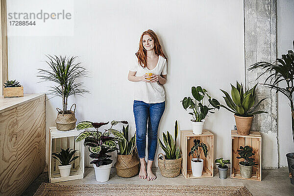 Lächelnde Frau mit einem Glas Saft inmitten von Zimmerpflanzen zu Hause