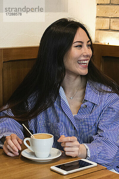 Lächelnde junge Frau mit Kaffeetasse  die wegschaut  während sie in einer Bar sitzt