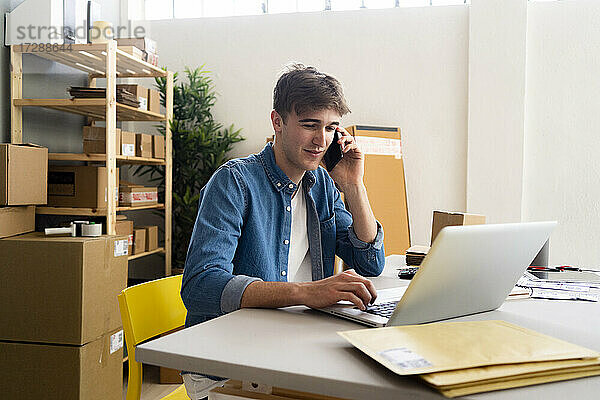 Männlicher Unternehmer  der einen Laptop benutzt  während er im Büro mit seinem Handy telefoniert