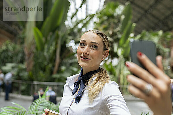 Junge Stewardess  die ein Selfie mit ihrem Smartphone im Garten eines Cafés macht