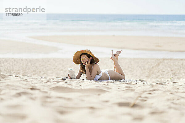 Junge Frau im Bikini liest ein Buch  während sie an einem sonnigen Tag am Strand im Sand liegt