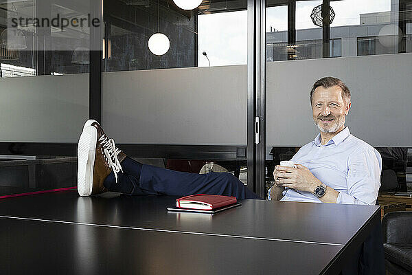 Lächelnde männliche Fachkraft mit hochgelegten Füßen  die einen Kaffee trinkt  während sie an einer Tischtennisplatte im Büro sitzt