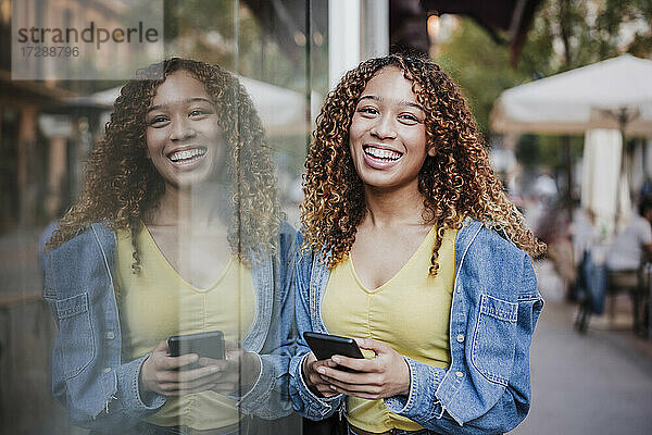 Lächelnde Frau mit Smartphone  die sich an eine Glaswand lehnt