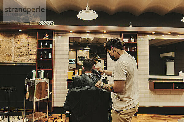Männlicher Friseur mit Gesichtsschutz beim Schneiden der Haare eines Kunden im Salon
