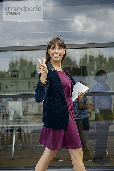 Lächelnde junge Geschäftsfrau mit digitalem Tablet und Friedenszeichen  die an einer Glaswand vorbeiläuft