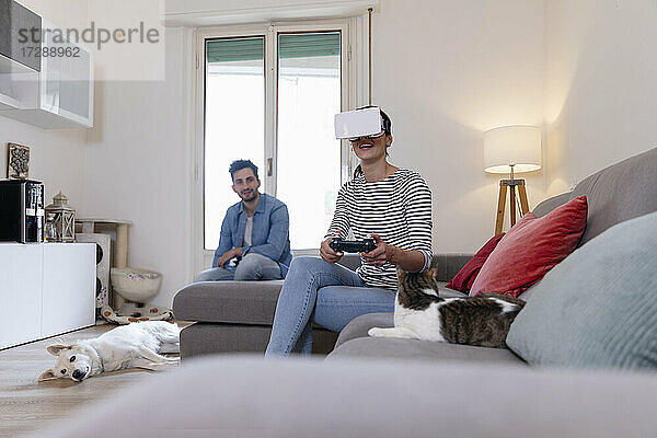 Junge Frau spielt mit virtueller Realität  während Mann und Haustiere zu Hause sitzen