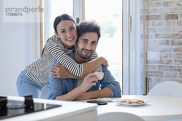 Lächelnde Freundin umarmt Freund mit Kaffeetasse in der Küche zu Hause