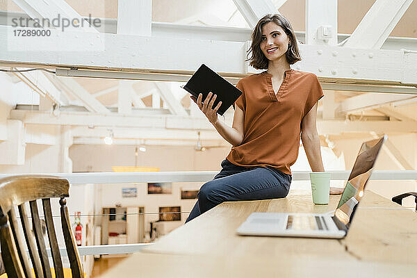 Lächelnde schöne Frau mit Tagebuch sitzt auf dem Schreibtisch im kreativen Büro und schaut weg