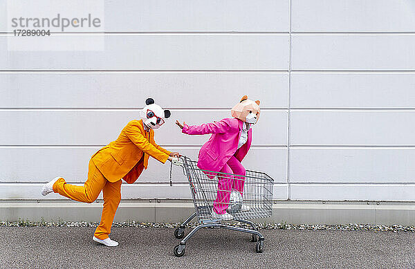 Mann und Frau in farbenfrohen Anzügen und Tiermasken  die mit einem Einkaufswagen herumspielen