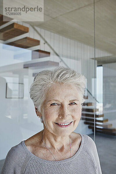 Lächelnde ältere Frau mit weißem Haar vor einer Glaswand stehend