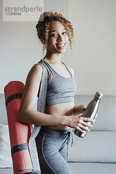 Junge Frau mit Gymnastikmatte und Flasche am Sofa im Wohnzimmer stehend