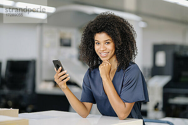 Lächelnde Geschäftsfrau mit Hand am Kinn  die ein Smartphone hält  während sie sich in der Industrie auf einen Tisch stützt