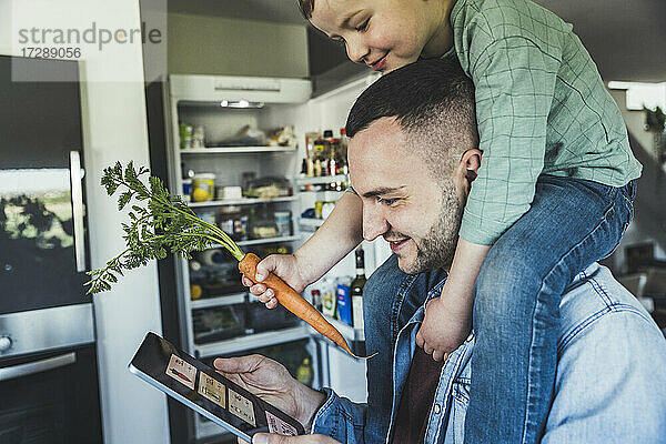 Lächelnder Mann  der ein Tablet benutzt  während sein Sohn in der Küche zu Hause auf der Schulter sitzt