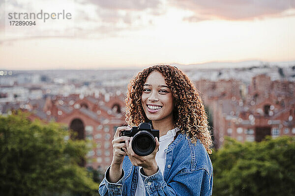Lächelnde Frau hält Kamera bei Sonnenuntergang