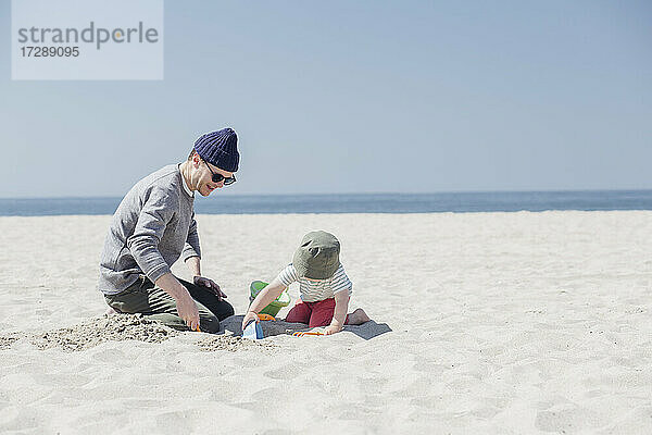Mann und Sohn spielen mit Sandspielzeug am Strand an einem sonnigen Tag