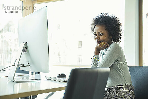Weibliche Fachkraft mit Hand am Kinn  die einen Computer auf dem Schreibtisch im Büro benutzt