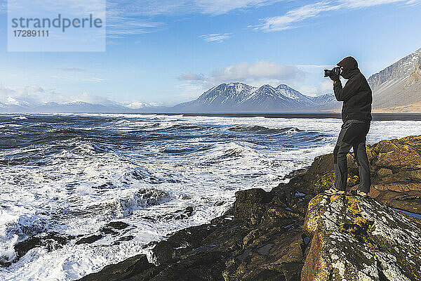 Älterer Mann steht auf einer Klippe und fotografiert das Meer