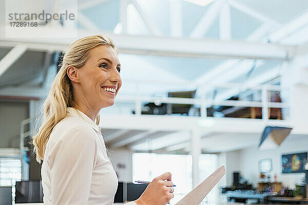 Lächelnde blonde weibliche Fachkraft mit Dokumenten in einem kreativen Büro schaut weg