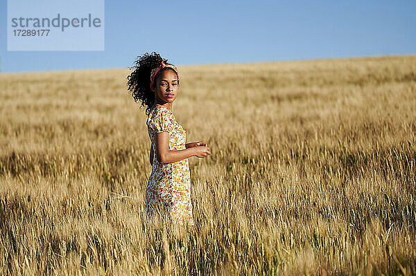Junge Frau mit lockigem Haar steht an einem sonnigen Tag in einem Weizenfeld