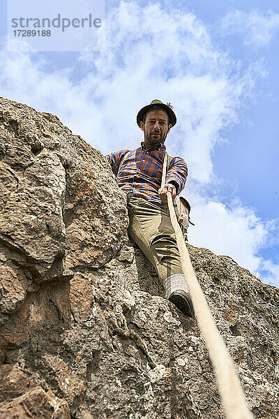 Männlicher Bergsteiger  der ein Seil hält  während er an einem sonnigen Tag auf einem Felsen sitzt