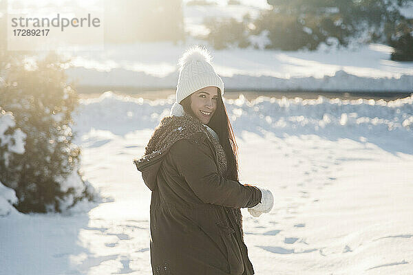 Lächelnde Frau schaut über die Schulter  während sie an einem sonnigen Tag im Schnee steht