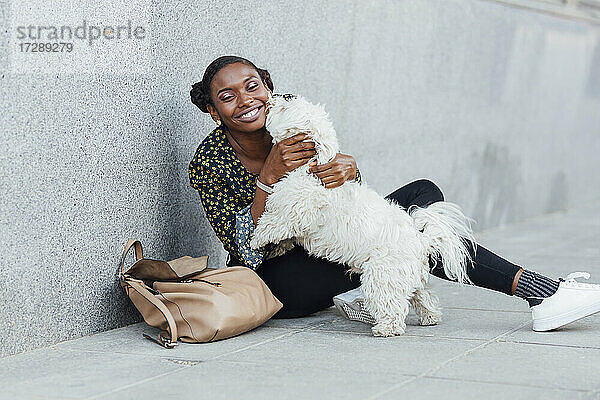 Lächelnde Frau  die mit ihrem Hund spielt  während sie auf dem Gehweg sitzt