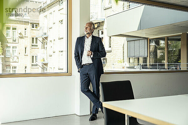 Männlicher Berufstätiger  der eine Kaffeetasse hält und sich an ein Glasfenster im Büro lehnt