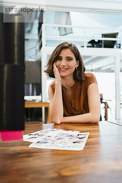 Lächelnde junge Frau  die mit der Hand am Kinn am Schreibtisch in einem kreativen Büro sitzt