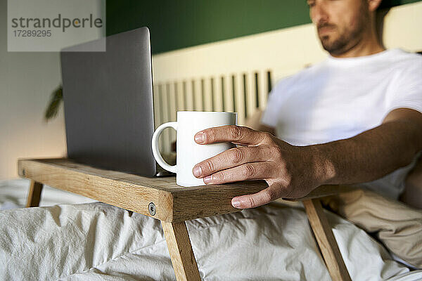 Mann arbeitet am Laptop  während er zu Hause im Bett sitzt
