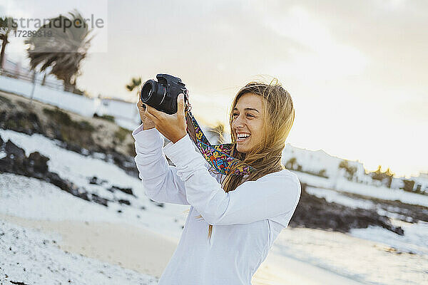 Lächelnde junge Frau beim Fotografieren durch die Kamera am Strand