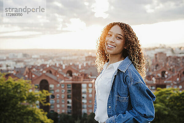 Lächelnde Frau in Jeansjacke in der Stadt bei Sonnenuntergang