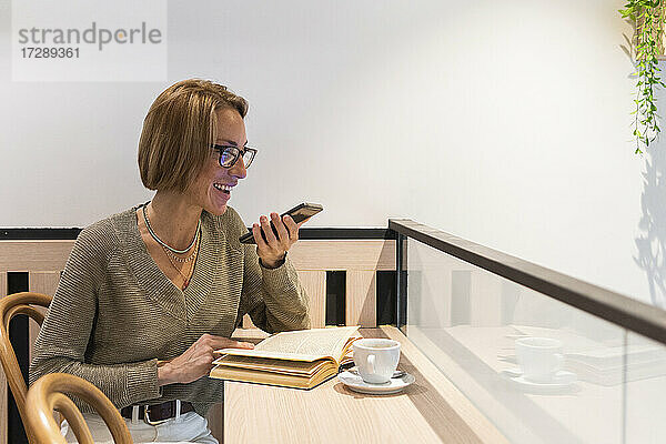 Lächelnde Frau mit Buch  die in einem Café mit ihrem Mobiltelefon spricht