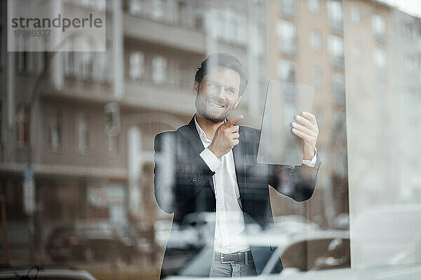 Lächelnder Geschäftsmann  der auf einen Videoanruf über ein digitales Tablet am Bürofenster zeigt
