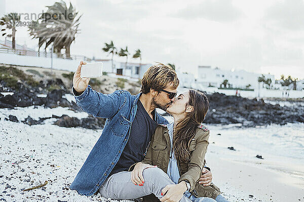 Freund küsst Freundin  während er ein Selfie mit dem Smartphone am Strand macht