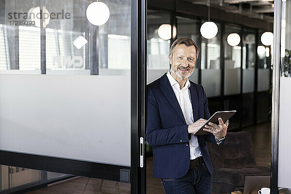Lächelnde männliche Fachkraft  die ein digitales Tablet hält und sich an eine Glastür im Büro lehnt