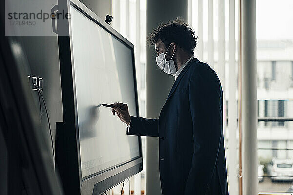 Männlicher Unternehmer mit Gesichtsmaske  der auf einer Projektionsfläche im Büro schreibt