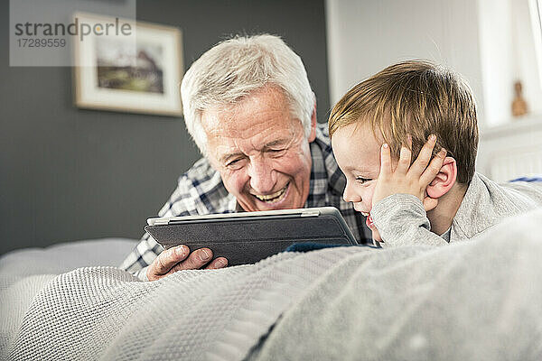 Lächelnder Großvater und Enkel benutzen ein digitales Tablet  während sie auf dem Bett liegen