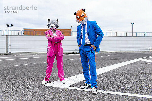 Mann und Frau in farbenfrohen Anzügen und Tiermasken posieren Seite an Seite auf einem leeren Parkplatz