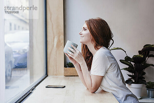 Lächelnde Frau  die einen Kaffee trinkt und sich zu Hause auf die Fensterbank lehnt