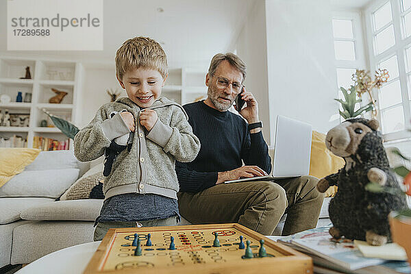 Lächelnder Junge  der ein Ludo-Spiel betrachtet  während sein Vater zu Hause im Hintergrund arbeitet