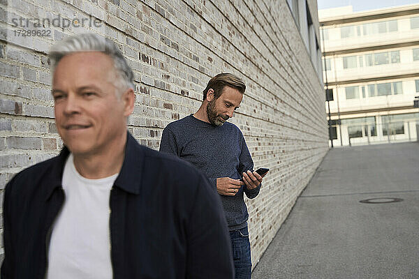 Männlicher Berufstätiger  der ein Mobiltelefon benutzt  während ein Geschäftsmann an der Wand vorbeiläuft