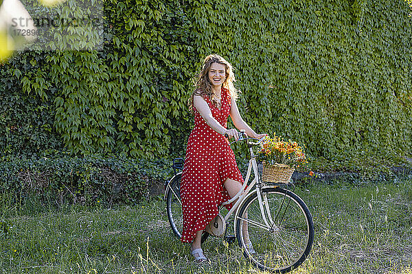 Fröhliche Frau auf dem Fahrrad sitzend an einer Efeuhecke im Garten
