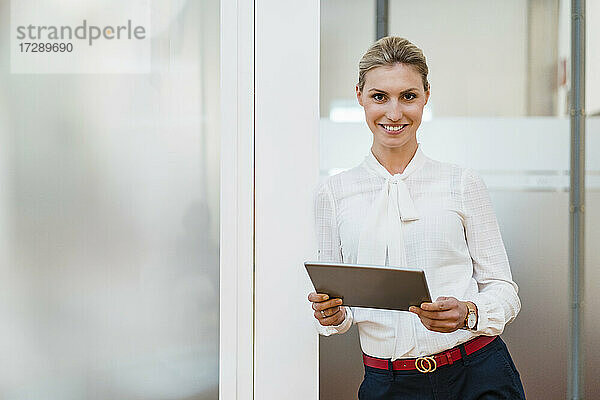 Lächelnde weibliche Fachkraft mit digitalem Tablet  die an der Tür im Büro lehnt