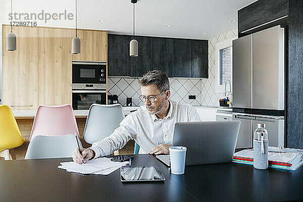 Männlicher Freiberufler mit Brille  der an einem Tisch im Heimbüro an Dokumenten schreibt