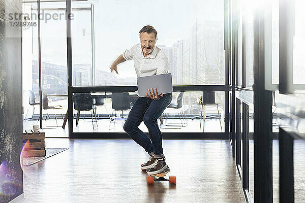 Älterer Geschäftsmann auf dem Skateboard mit Blick auf den Laptop im Büro